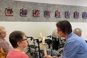 Eine Person zündet eine Kerze für eine Seniorin in einem Raum mit Sitzmöglichkeiten und Bildern an der Wand.