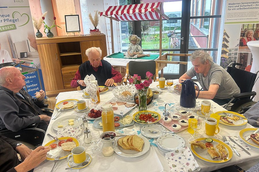 Mehrere ältere Menschen sitzen an einem runden Tisch mit weißer Tischdecke und gedecktem Frühstück mit Brot, Aufschnitt, Getränken und Blumen in einem hellen Raum