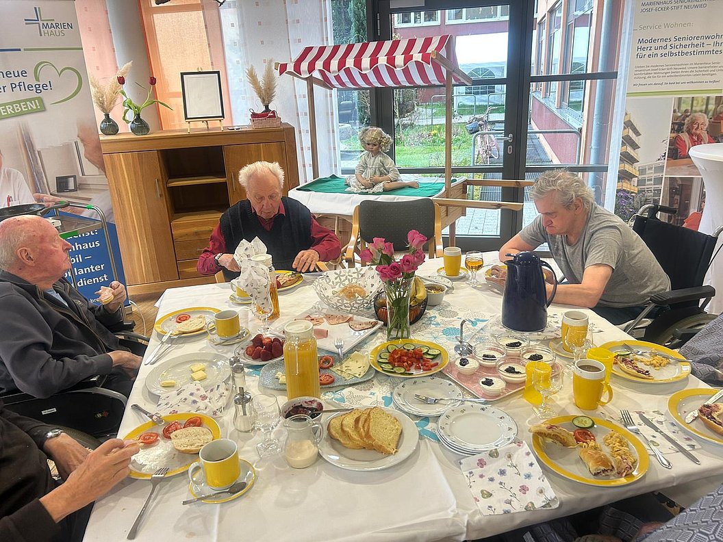 Mehrere ältere Menschen sitzen an einem runden Tisch mit weißer Tischdecke und gedecktem Frühstück mit Brot, Aufschnitt, Getränken und Blumen in einem hellen Raum