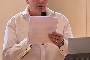 Älterer Mann mit Brille liest von einem Blatt vor einem Mikrofon und einem Rednerpult