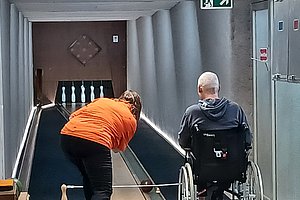 Person in oranger Jacke kniet vor Person im Rollstuhl in einem langen, schmalen Flur mit grauen Wänden