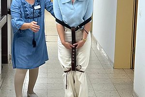 Frau mit blauer Bluse und weißer Hose trägt eine Gehhilfe mit Gurten, eine andere Frau in blauem Kleid begleitet sie im Flur.