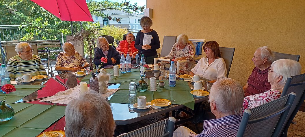 Gruppe älterer Menschen sitzt an einem großen Tisch im Freien unter einem roten Sonnenschirm und trinkt Kaffee