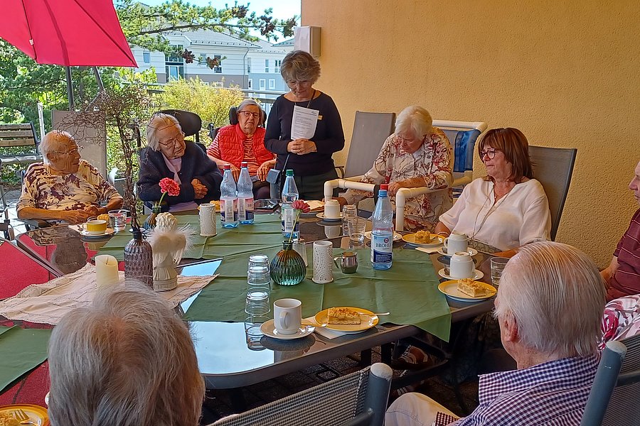 Gruppe älterer Menschen sitzt an einem großen Tisch im Freien unter einem roten Sonnenschirm und trinkt Kaffee