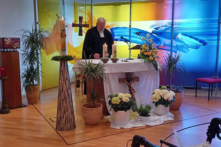 Ein Priester steht an einem mit Kerzen und Blumen geschmückten Altar in einer Kirche mit farbigem Glasfenster im Hintergrund, während mehrere Personen auf Stühlen sitzen.