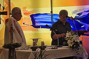 Zwei Personen stehen an einem Altar mit Kerzen und Blumen vor einer farbigen Wand mit abstrakten Mustern