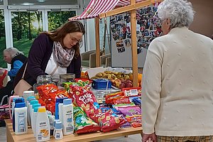 Markttisch mit Lebensmitteln und Pflegeprodukten, zwei Personen stehen davor und schauen das Angebot an.