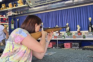 Frau schießt mit einem Luftgewehr auf eine Kirmesattraktion, im Hintergrund bunte Dekoration.