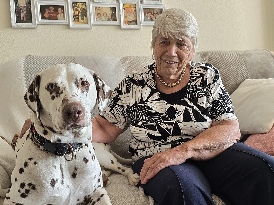 Seniorin mit grauen Haaren, sitzt auf einem Sofa neben einem süßen Dalmatinerhund, beide lächeln.