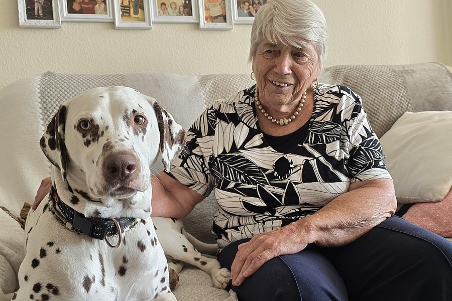 Seniorin mit grauen Haaren, sitzt auf einem Sofa neben einem süßen Dalmatinerhund, beide lächeln.