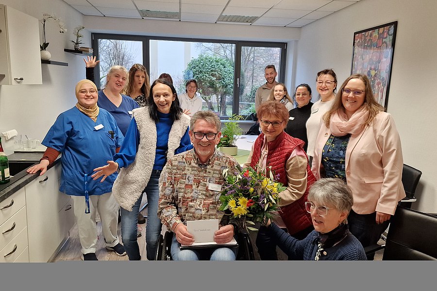 Gruppe von Menschen in einem hellen Raum mit Küchenzeile, eine Person im Rollstuhl hält ein Buch, eine andere überreicht einen Blumenstrauß.
