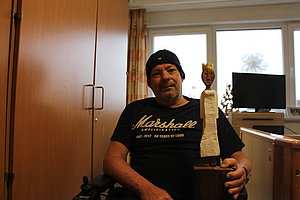 Mann im Rollstuhl hält eine Holzfigur in der Hand, im Hintergrund ein heller Raum mit Möbeln.