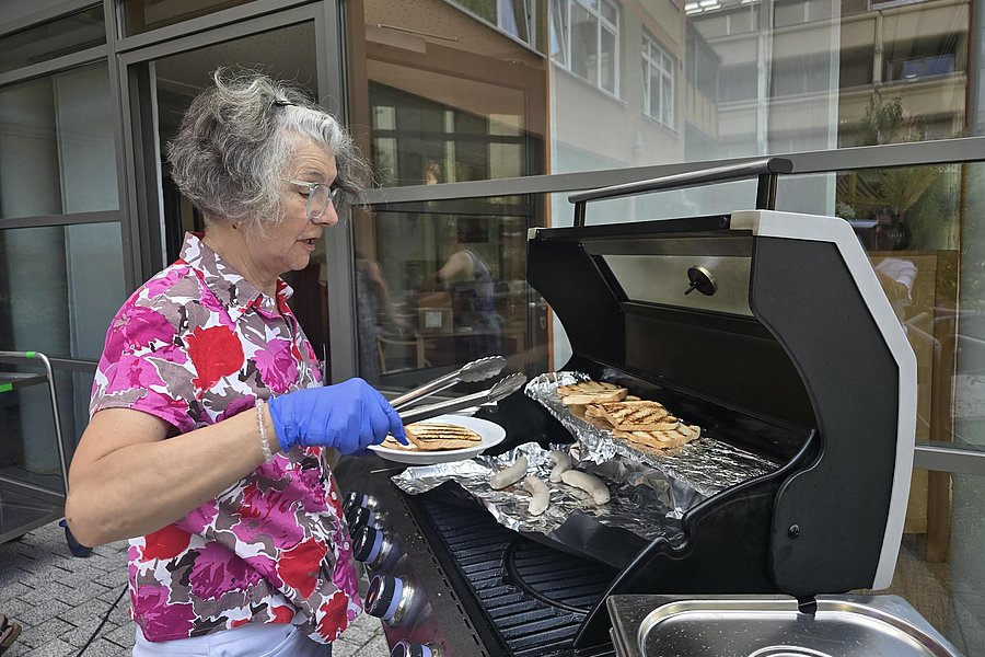 Ältere Frau mit grauen Haaren und blauem Handschuh wendet mit Zange Fischfilets auf einem Gasgrill auf einer Terrasse vor einem Gebäude
