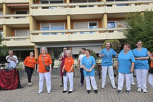 Gruppe von Menschen in orangefarbenen und hellblauen Arbeitskleidungen steht vor einem mehrstöckigen Wohngebäude im Freien