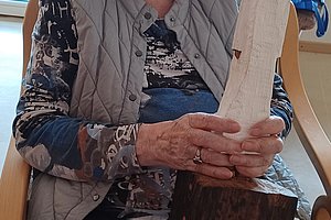 Ältere Person sitzt auf einem Stuhl und hält eine geschnitzte Holzfigur in den Händen