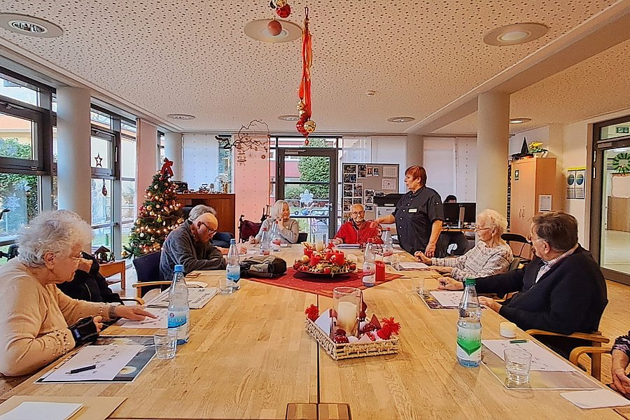 Mehrere ältere Menschen sitzen an einem langen Holztisch in einem hellen Raum mit Weihnachtsdekoration und sprechen miteinander.