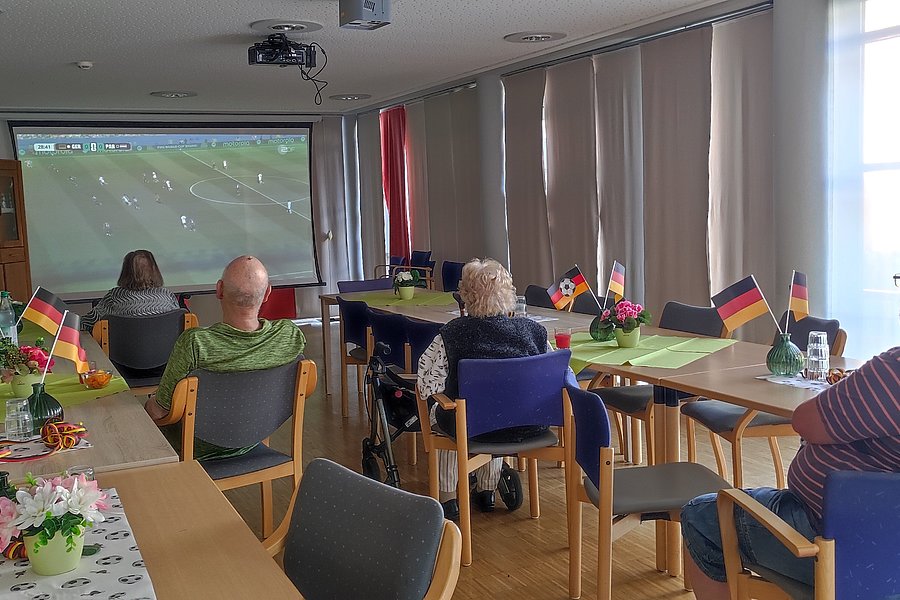 Mehrere ältere Menschen sitzen in einem Raum mit Tischen, auf denen kleine deutsche Fahnen stehen, und schauen auf eine Leinwand mit einem Fußballspiel.