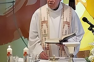 Priester in weißem Gewand steht hinter einem Altar mit Kerzen, Kelch und Buch vor einem Wandbild mit Kreuz