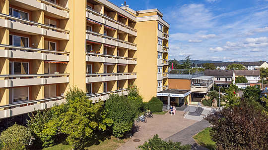 Mehrstöckiges Wohngebäude mit Balkonen, davor ein gepflasterter Weg und mehrere Bäume unter blauem Himmel