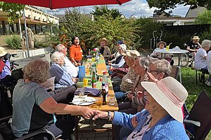 Gruppe älterer Menschen sitzt an einem langen Tisch im Freien unter einem großen roten Sonnenschirm und hält sich an den Händen
