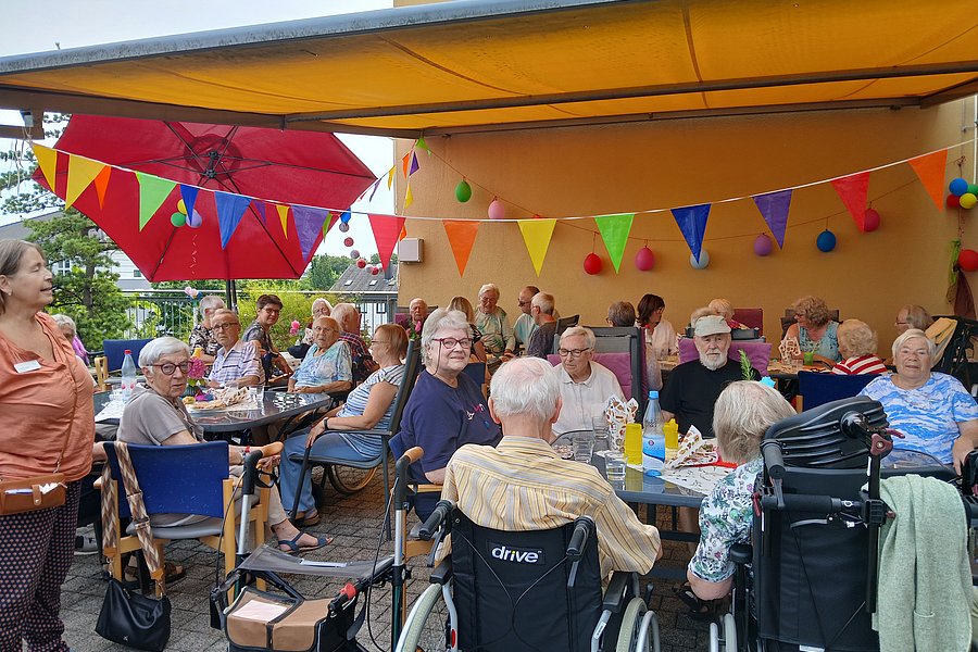 Gruppe älterer Menschen sitzt an mehreren Tischen unter einem gelben Sonnensegel mit bunten Wimpeln und Luftballons, einige im Rollstuhl, im Außenbereich eines Gebäudes