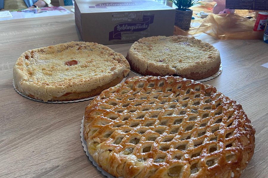 Drei verschiedene Kuchen auf einem Holztisch, im Hintergrund eine weiße Verpackung mit der Aufschrift 'Lieblingsstücke' und eine grüne Pflanze