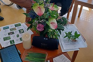 Blumenstrauß auf einem Tisch mit verstreuten Papieren, einem Lautsprecher und einem Tablet, umgeben von Personen