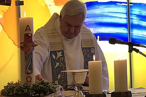 Ein Priester in weißem Gewand mit blauem Stola steht vor einem Altar mit Kerzen, Kelch und einem großen Osterkerze mit Alpha- und Omega-Symbolen