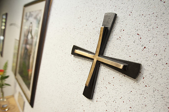 Metallkreuz mit breiten Enden an der Wand, unscharf im Hintergrund ein gerahmtes Bild und Pflanzen.