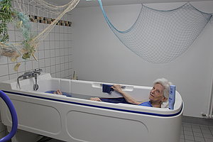 Seniorin in einer modernen Badewanne, entspannt beim Baden mit Handtuch. Helle Badezimmeratmosphäre.