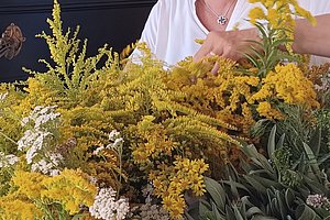 Person sitzt hinter einem großen Strauß aus verschiedenen gelben und weißen Blumen und grünen Blättern in einem hellen Raum mit weißer Wand und einem gerahmten Bild an der Wand
