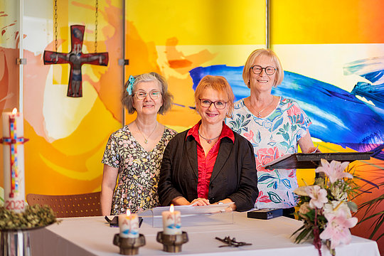Drei Frauen stehen hinter einem Altar mit brennenden Kerzen, Blumen und einem Kreuz an der gelb-blau bemalten Wand.