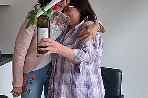 Zwei Frauen umarmen sich, eine hält eine Flasche Wein mit Blumen.Arrangement, freundliche Atmosphäre.