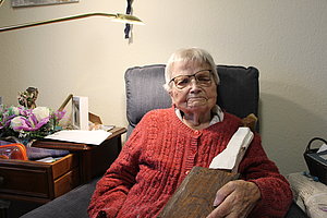 Sitzende ältere Dame in rotem Pullover, lächelnd, mit Holzstück und Blumen im Hintergrund.