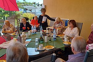 Gruppe älterer Menschen sitzt an einem großen Tisch im Freien unter einem roten Sonnenschirm und trinkt Kaffee