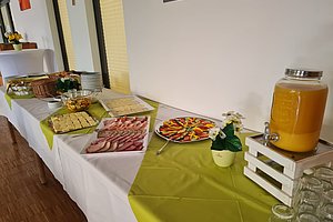 Buffet mit verschiedenen kalten Speisen, einem Getränkespender mit orangem Saft und Gläsern auf einem mit grünem Tischtuch bedeckten Tisch in einem Raum mit Holzboden