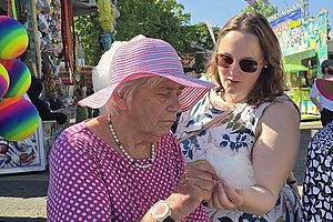 Ältere Frau mit rosa Hut und gepunktetem Kleid zeigt etwas auf einem Smartphone einer jüngeren Frau in einem geblümten Kleid auf einem Jahrmarkt