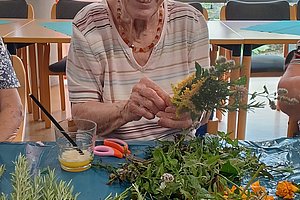 Ältere Frau sitzt an einem Tisch mit verschiedenen frischen Kräutern und Blumen und hält einen Strauß in den Händen