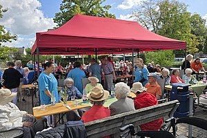 Menschen sitzen und stehen unter einem roten Pavillon im Freien bei einer Veranstaltung an Holztischen und Bänken