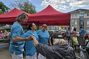 Drei Personen in blauen T-Shirts stehen vor einem roten Zelt und halten die Hand einer im Rollstuhl sitzenden Person