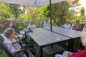 Mehrere ältere Menschen sitzen im Garten an einem großen rechteckigen Tisch unter einem Sonnenschirm und unterhalten sich.