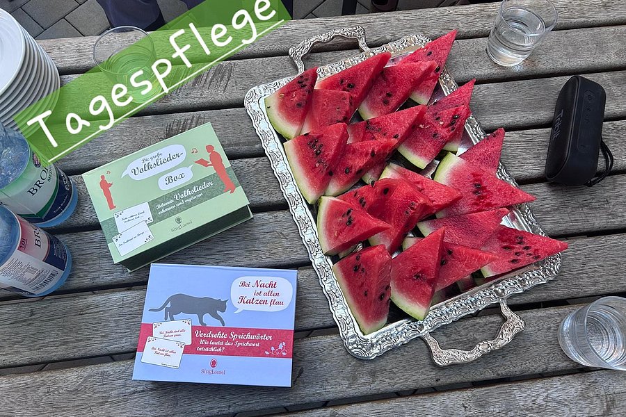 Auf einem Holztisch steht eine silberne Platte mit dreieckigen Wassermelonenstücken, daneben liegen zwei bunte Kartons und mehrere Gläser mit Wasser.