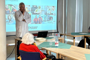 Ein Mann in weißem Kittel steht vor einem Beamerbild mit Fotos und Text, vor ihm sitzt eine ältere Person im Rollstuhl in einem Raum mit Tischen und Stühlen.