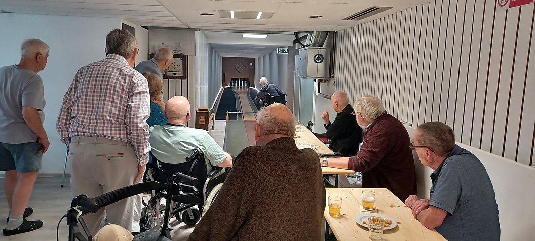Gruppe älterer Männer sitzt und steht an einem langen Tisch in einem Raum mit Kegelbahn, ein Mann kegelt gerade