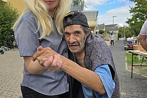 Älterer Mann mit Baseballkappe tanzt mit jüngerer Frau mit langen blonden Haaren im Freien auf gepflastertem Platz.