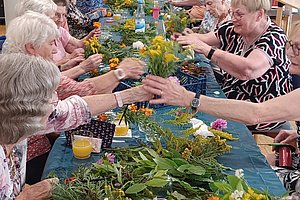 Seniorengruppe bei einem kreativen Workshop mit Blumen und Pflanzen auf einem langen Tisch.