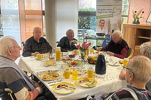 Sechs ältere Menschen, zwei im Rollstuhl, sitzen an einem gedeckten Tisch in einem hellen Raum und essen gemeinsam.
