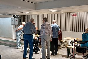 Gruppe älterer Menschen steht und sitzt an einem langen Tisch in einem hellen Raum mit weißer Wandverkleidung