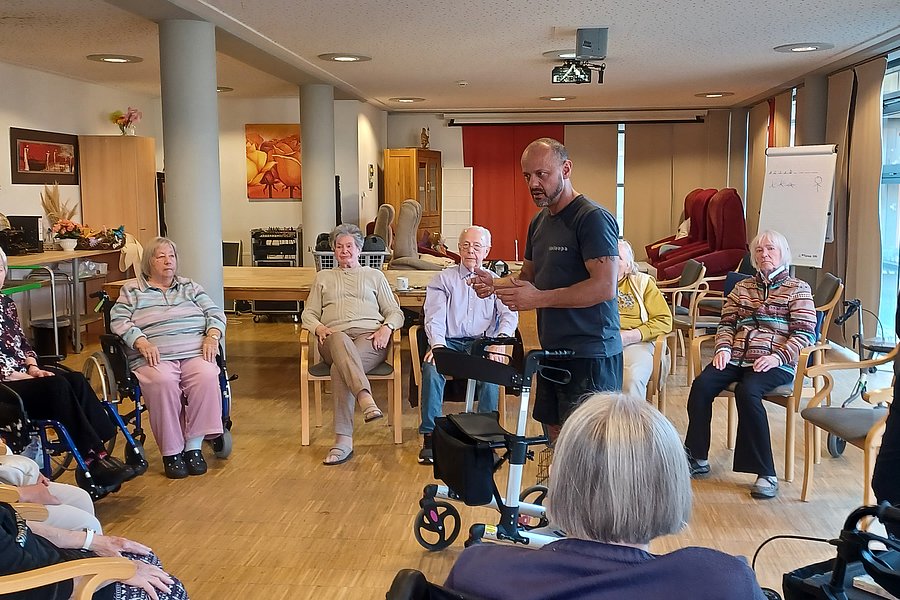 Mehrere ältere Menschen sitzen in einem Stuhlkreis in einem hellen Raum, zwei Männer stehen in der Mitte und sprechen zu der Gruppe.