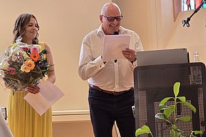 Mann mit Brille liest von einem Blatt vor einem Mikrofon stehend neben einer Frau in gelbem Kleid mit Blumenstrauß in einem hellen Raum